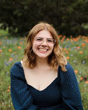 Smiling woman with wavy auburn hair and clear glasses wearing a navy top in a field of orange and purple wildflowers.