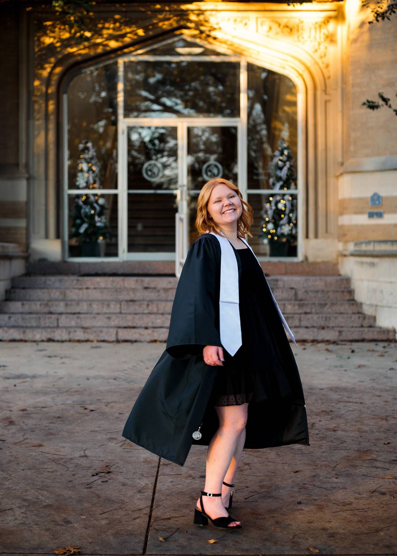A smiling graduate in a black gown and white stole poses on stone steps in front of a historic campus building.