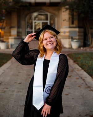 A smiling graduate in a black cap and white stole tips her cap on a brick pathway in front of a campus building.