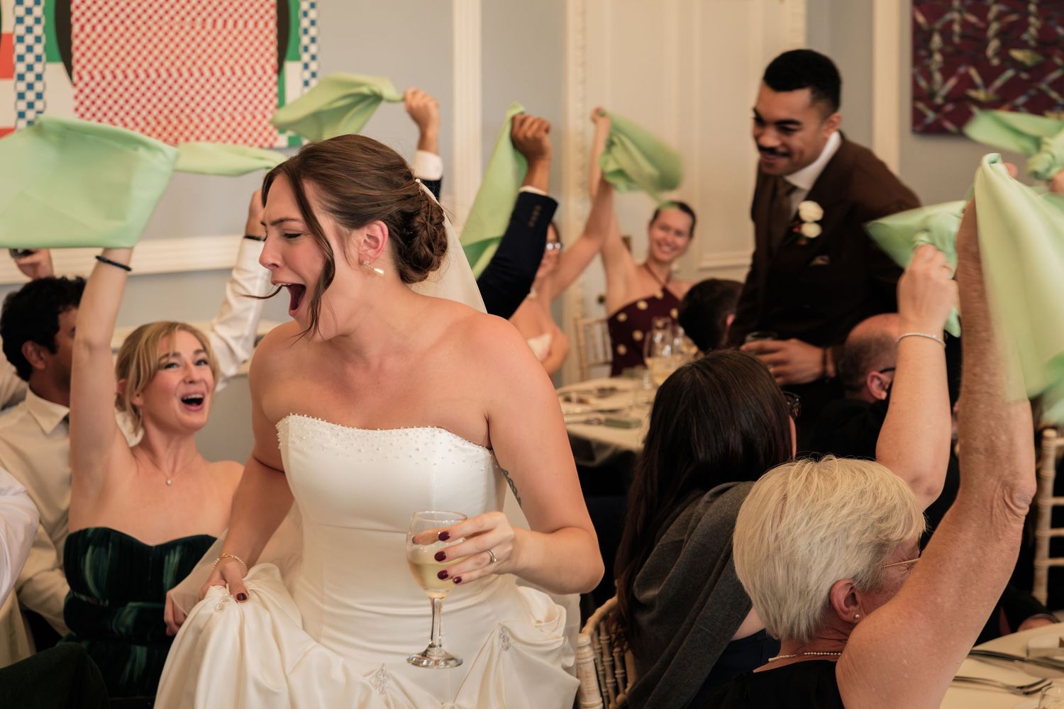 Bride in a white strapless gown holds a champagne glass while wedding guests wave green napkins and cheer at the reception.