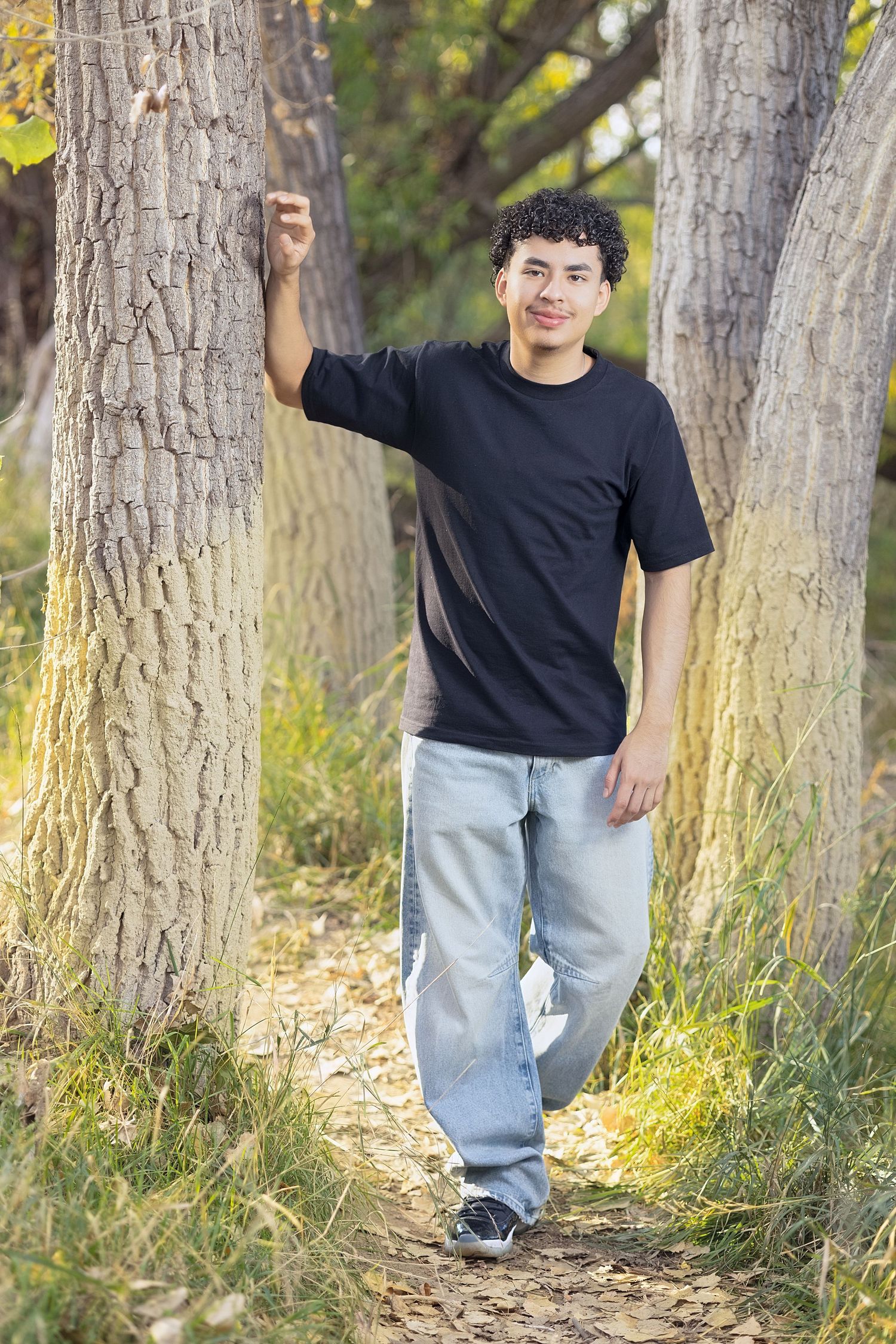 Young man with curly hair sits cross-legged on a wooden bridge, smiling, wearing a black t-shirt and jeans.