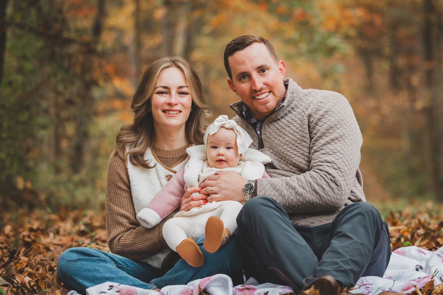Smiling family of three sits on a fall blanket in an autumn forest, parents holding their baby girl in a white bow headband in Lake Fairfax.
