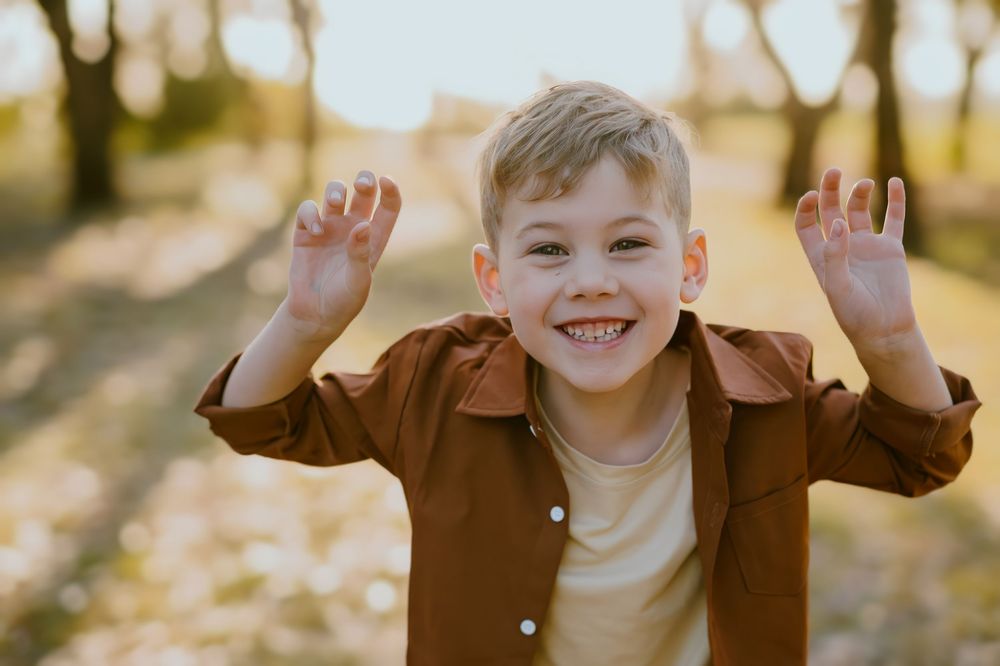 A cheerful young blonde boy in a brown shirt raises his hands outdoors in golden sunlight.
