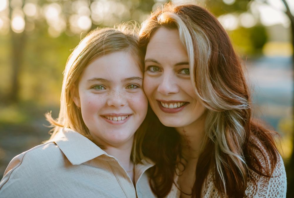 Two smiling women pose cheek to cheek outdoors in warm golden hour light, sharing a joyful close-up portrait.