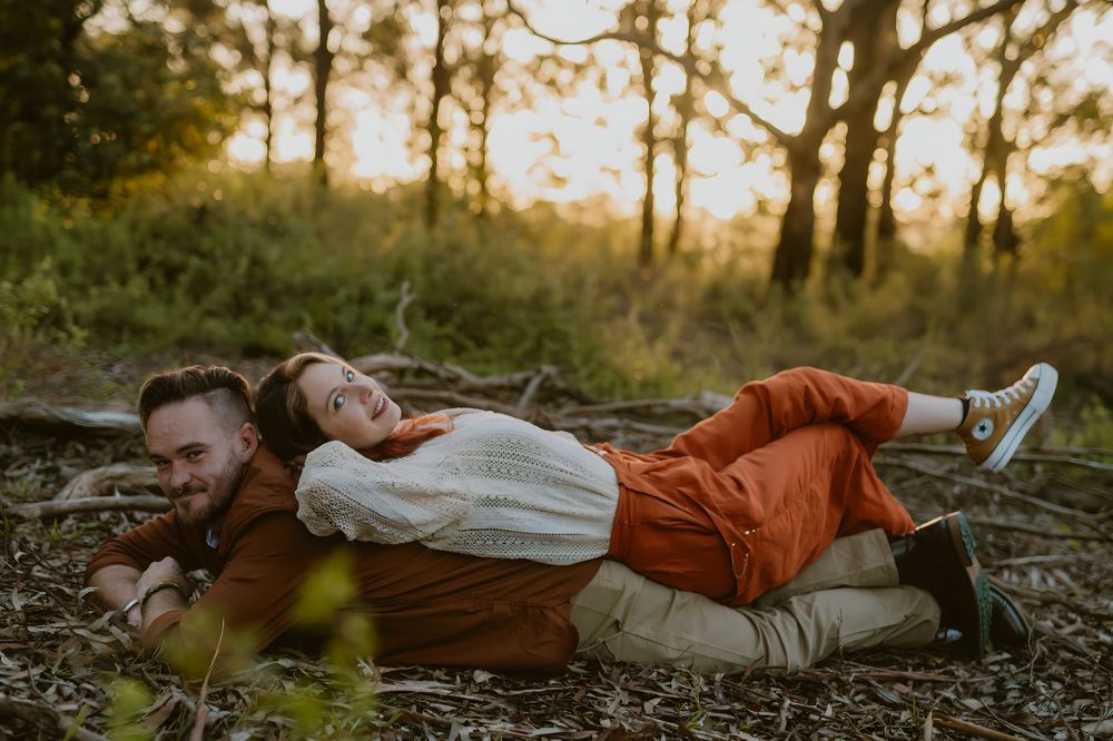 Couple laughing and lying together on forest floor during golden hour sunset photography session.