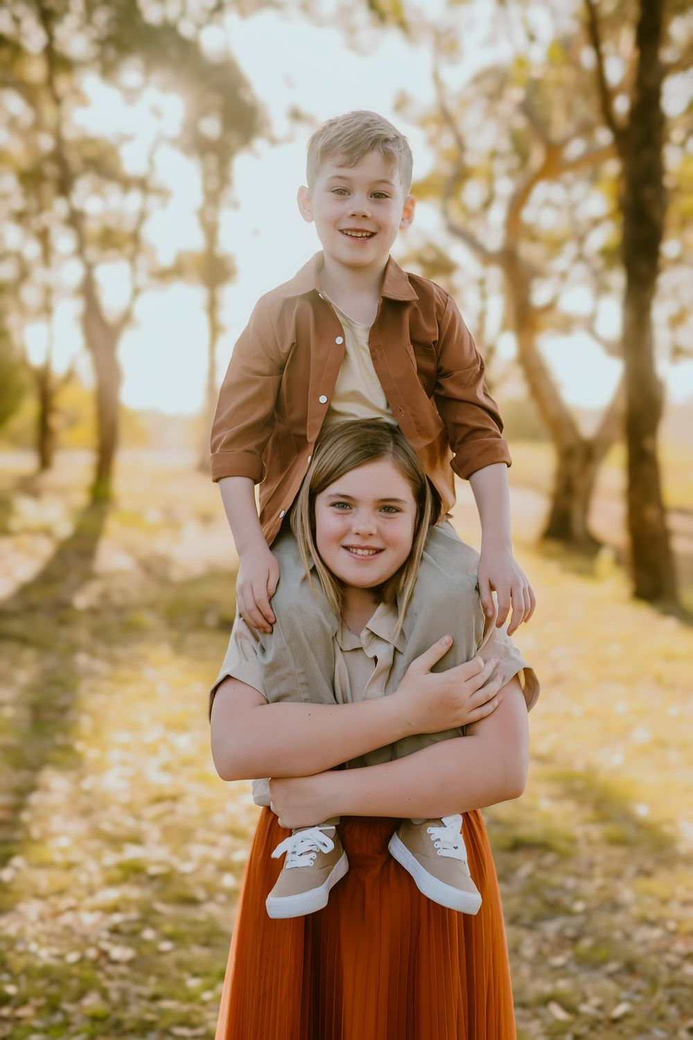 Two smiling children pose outdoors at golden hour, with a boy on a girl's shoulders among sunlit trees.