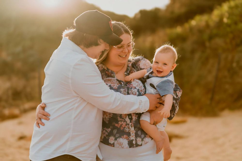 Family with baby enjoying golden sunset at beach, parents smiling and embracing newborn in floral setting.