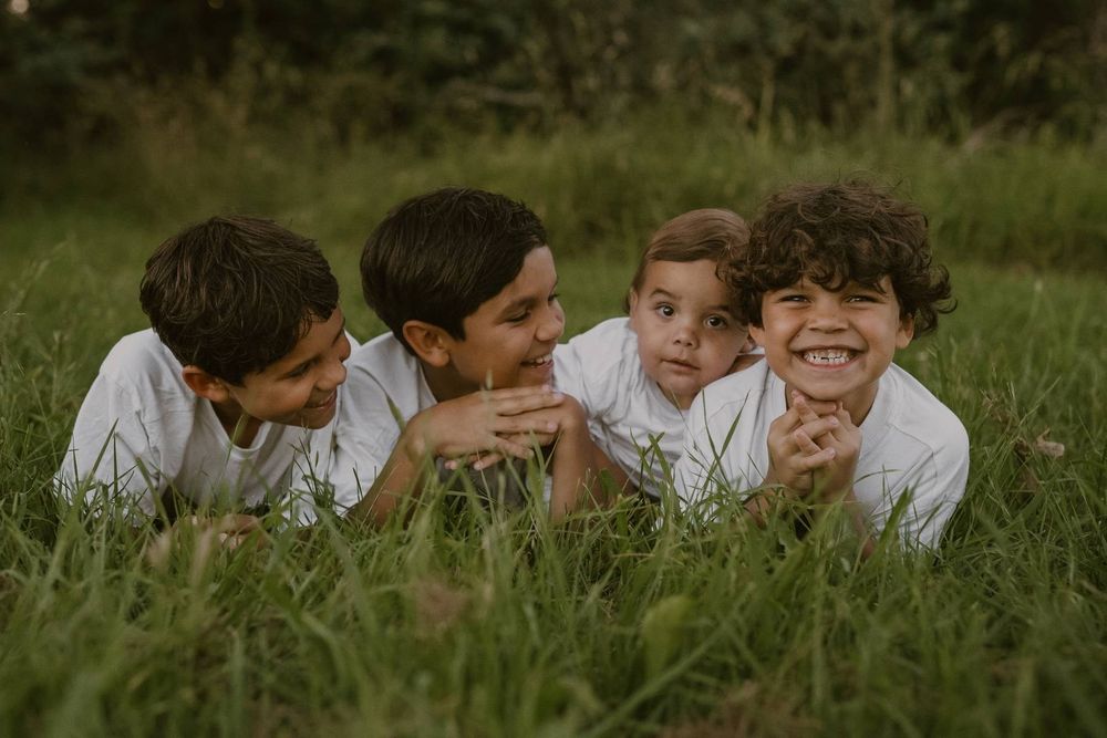Four boys in white shirts lying in green grass, smiling and laughing together outdoors.