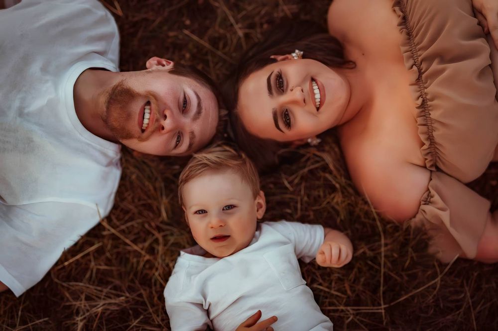 Happy family lying on grass, two smiling parents with their baby between them, aerial view outdoor photo.