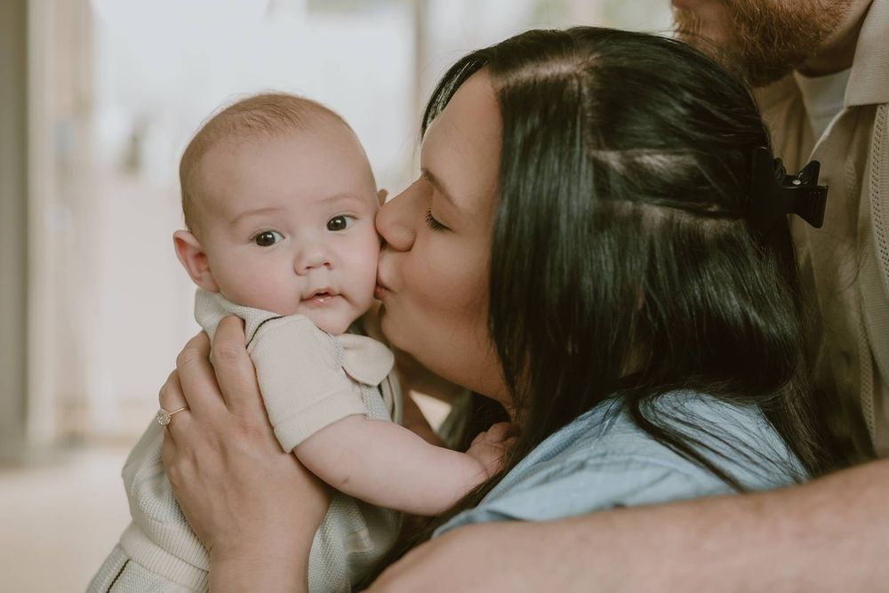 Loving mother kissing her newborn baby on the cheek while father holds the infant gently indoors.