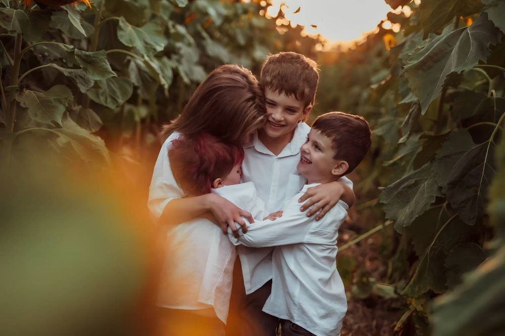 A mother hugs her two young sons in a sunflower garden during golden hour, all wearing white shirts.