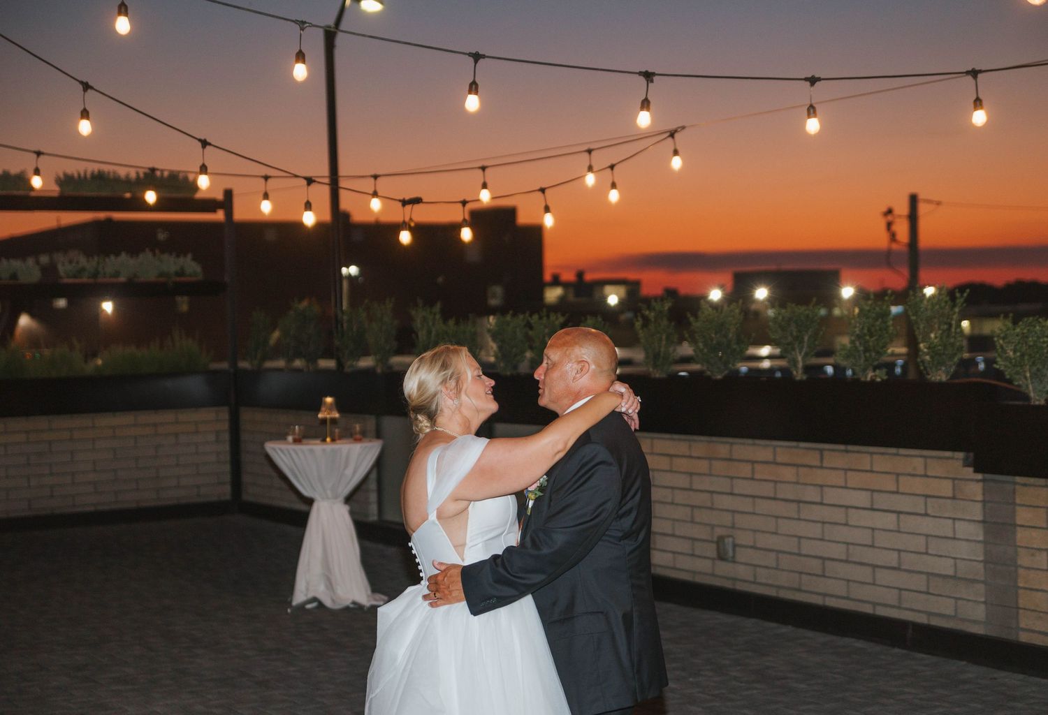 Bride and groom share first dance on rooftop venue under string lights at sunset.