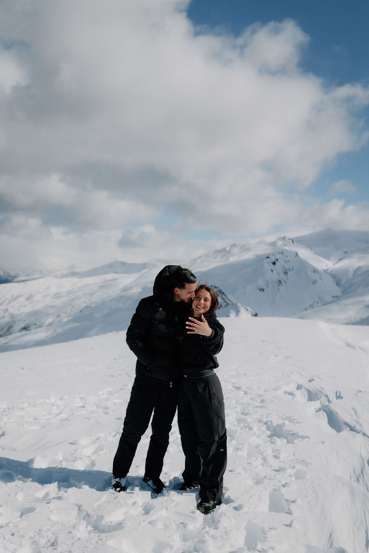 Couple embracing and smiling on a snowy mountain peak, wearing black winter gear with cloudy blue skies behind them.