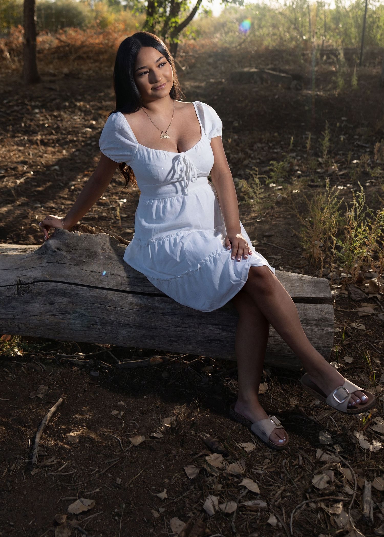 Young woman in white dress sitting on a log in a natural outdoor setting, smiling warmly at the camera.
