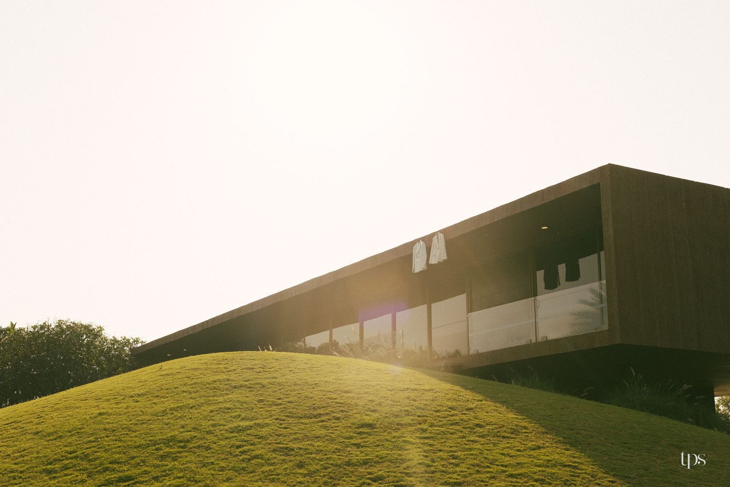 Modern elongated building with large glass windows and dark cladding rests atop a grassy green hill, glowing in warm golden sunlight.