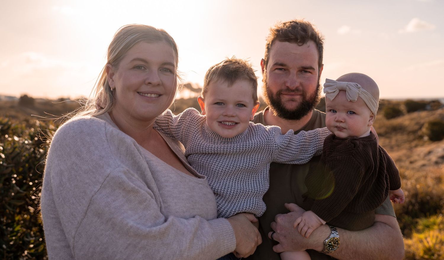 Happy family of four posing outdoors at golden hour, parents holding two young children in a scenic natural setting.