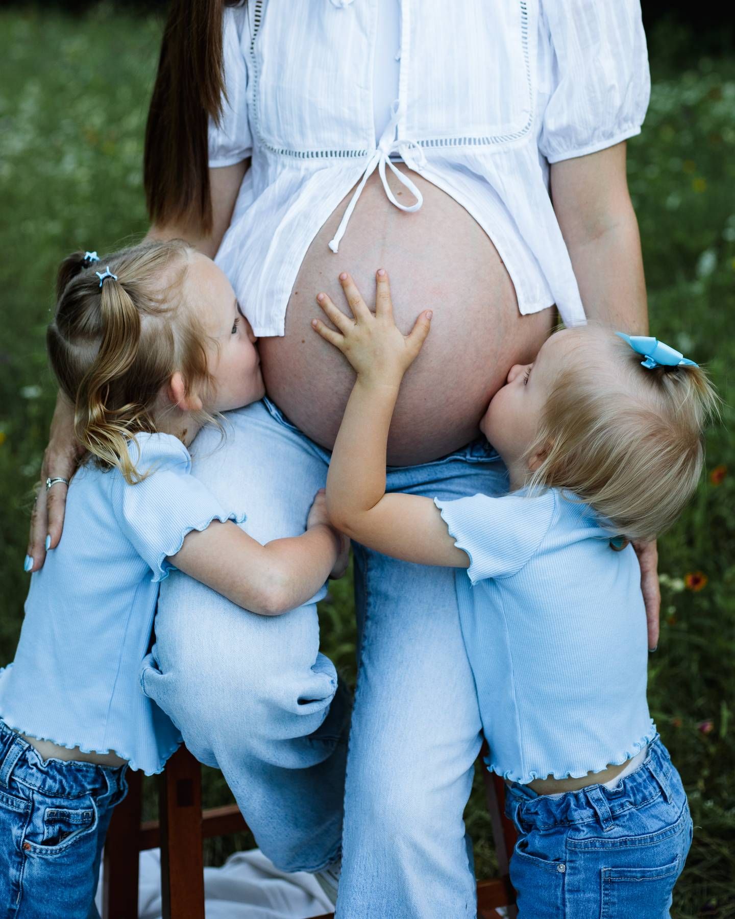 Two toddler girls in matching light blue outfits lovingly hug and kiss their expectant mother's bare pregnant belly outdoors.