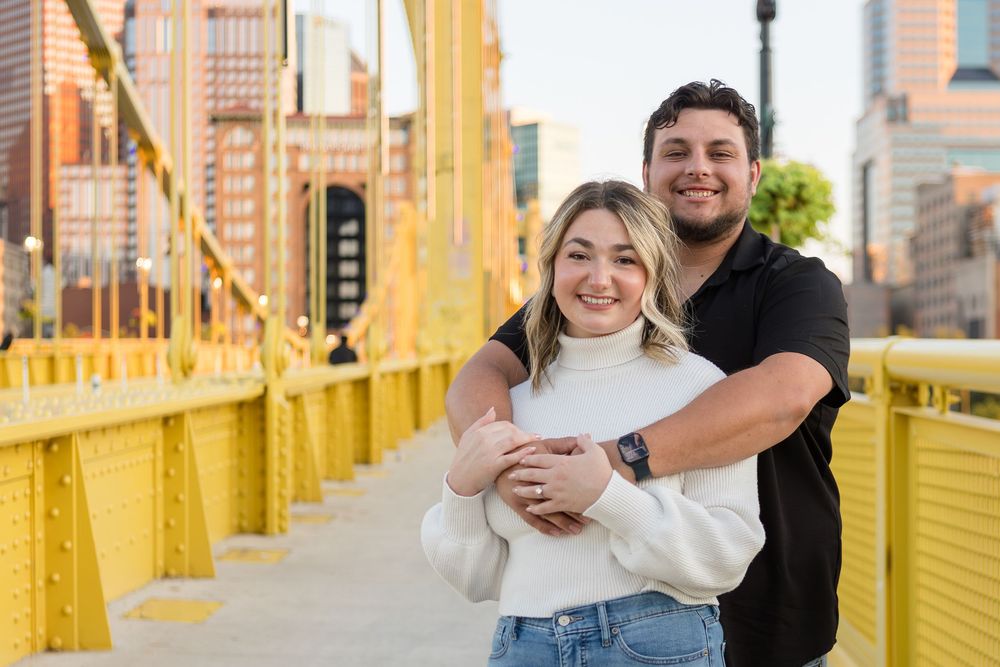 Smiling couple embracing during an engagement photo session on a yellow steel bridge with a city skyline in the background.