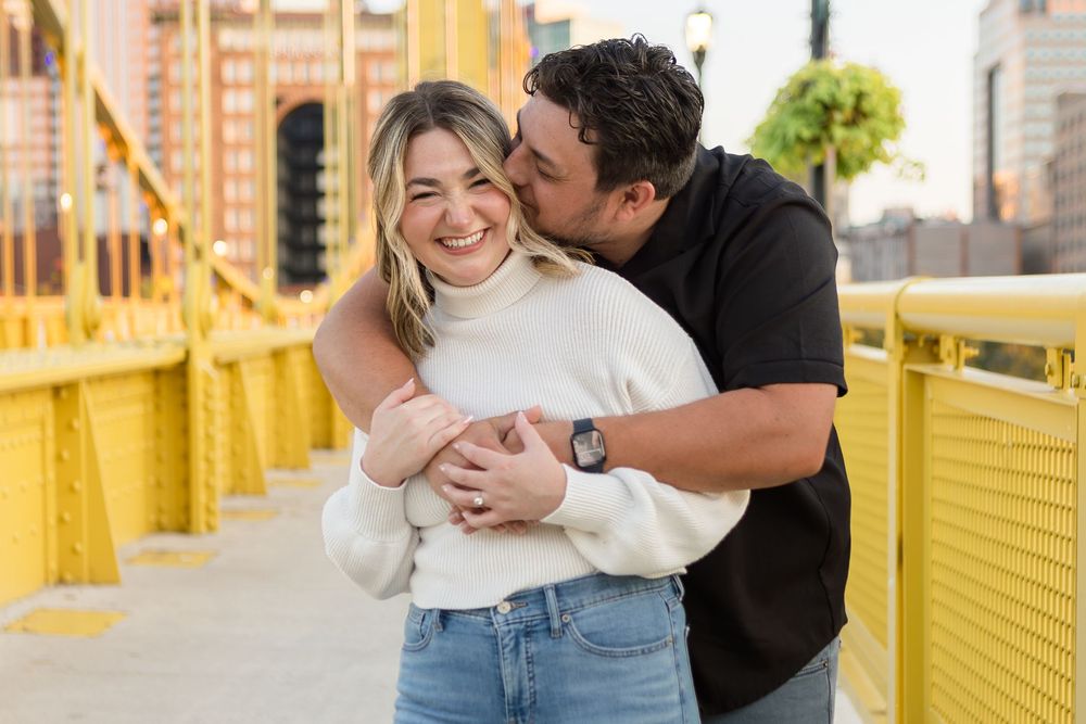 Smiling woman in a white sweater and jeans is embraced and kissed on the cheek by a man in a black shirt on a yellow bridge.