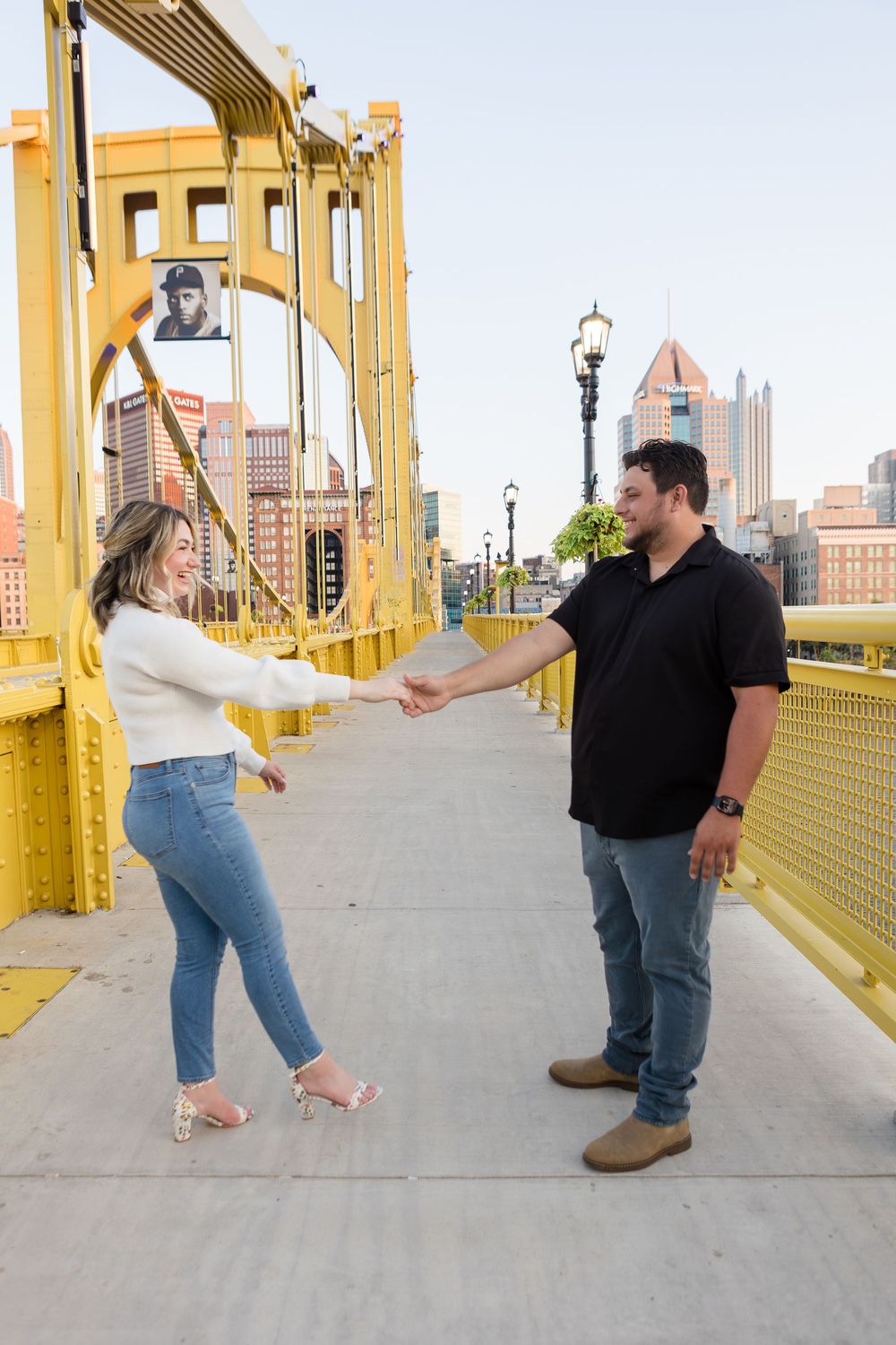 Couple holding hands and facing each other on a yellow suspension bridge with a city skyline and vintage street lamps behind them.