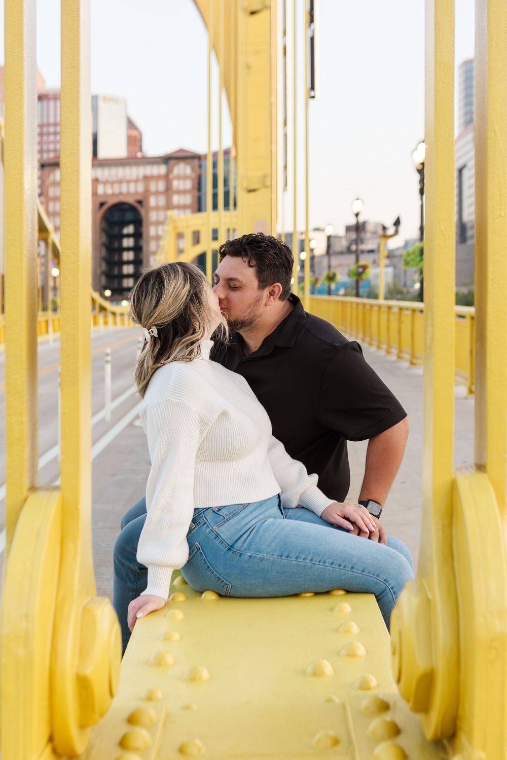 Couple kissing while sitting on a yellow steel bridge railing with a city skyline in the background during an engagement photo session.