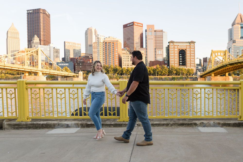 Couple holding hands and walking along a yellow-railed bridge with a sunny city skyline in the background.