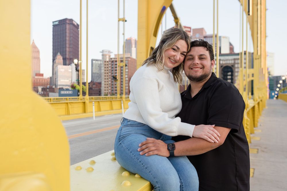 Smiling couple in white sweater and black shirt embrace on a yellow steel bridge with a city skyline behind them.