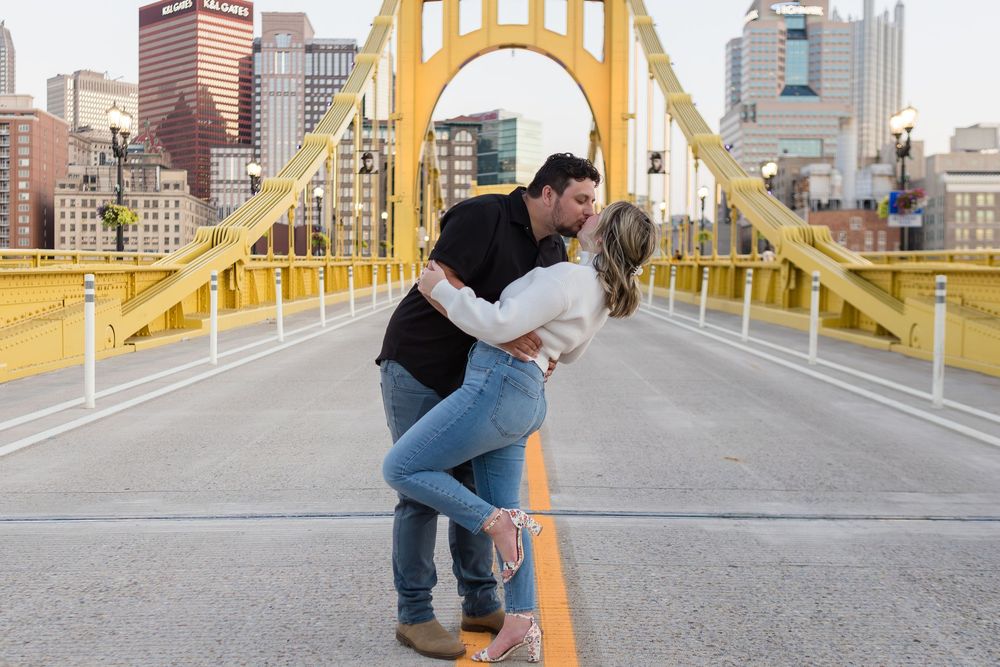 Couple sharing a romantic dip kiss on a yellow suspension bridge with a city skyline in the background during an engagement photo session.