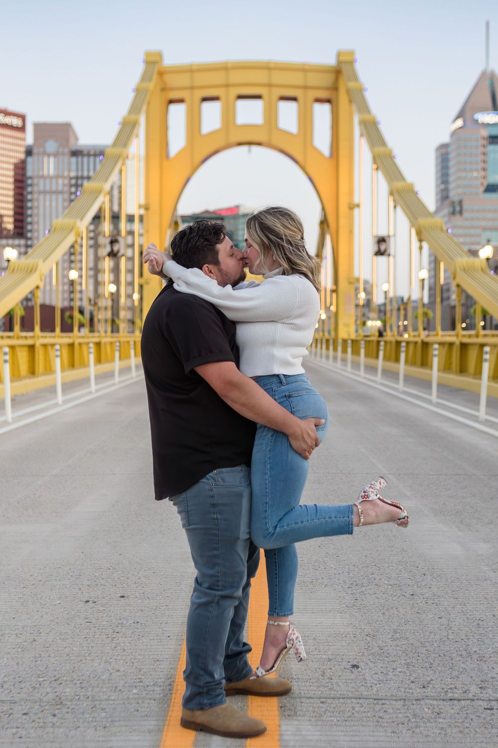 Couple embracing and kissing on a yellow suspension bridge with city buildings in the background during an engagement photo session.