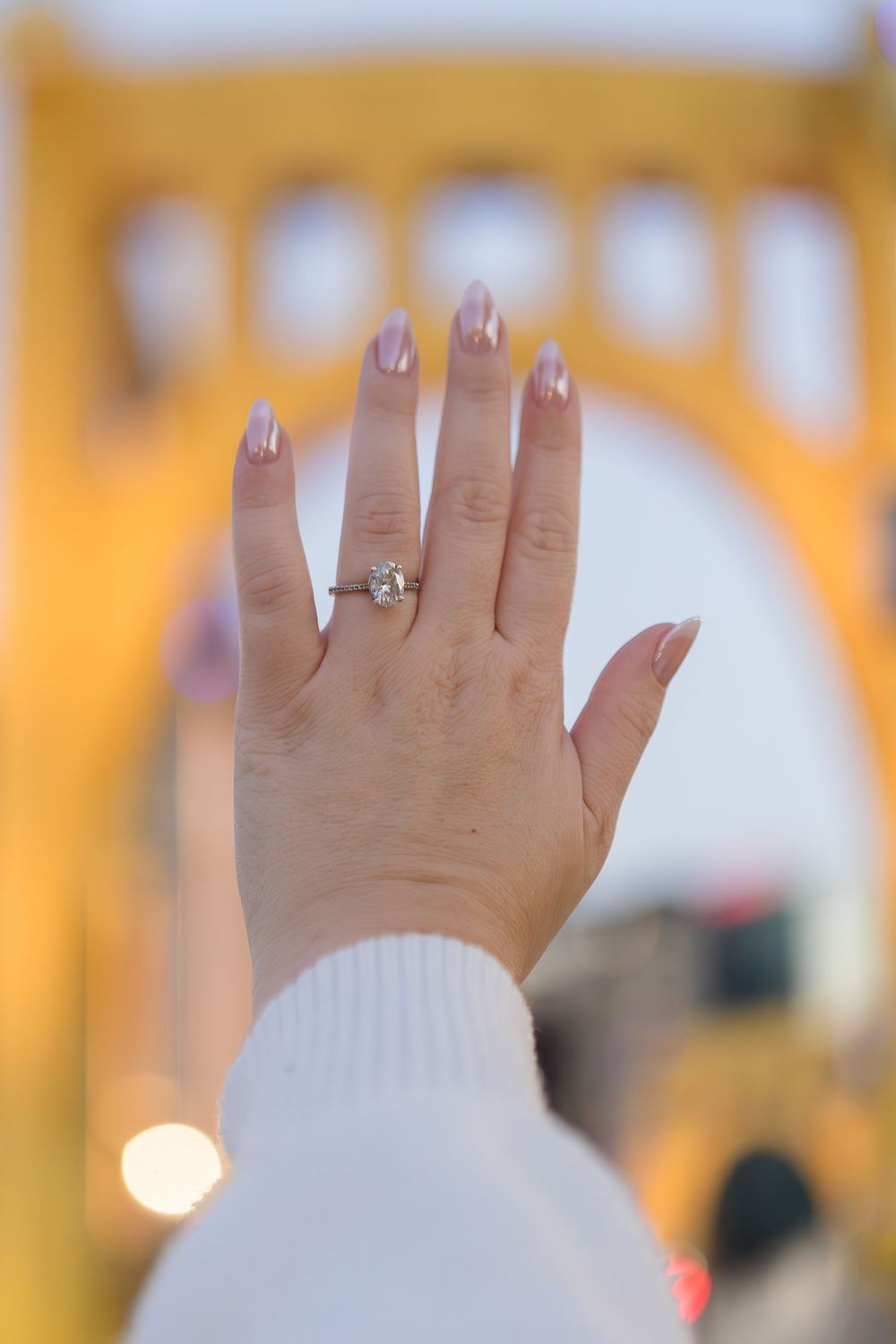 Hand with pale pink manicure raised to show a diamond engagement ring against a blurred golden yellow bridge at dusk.