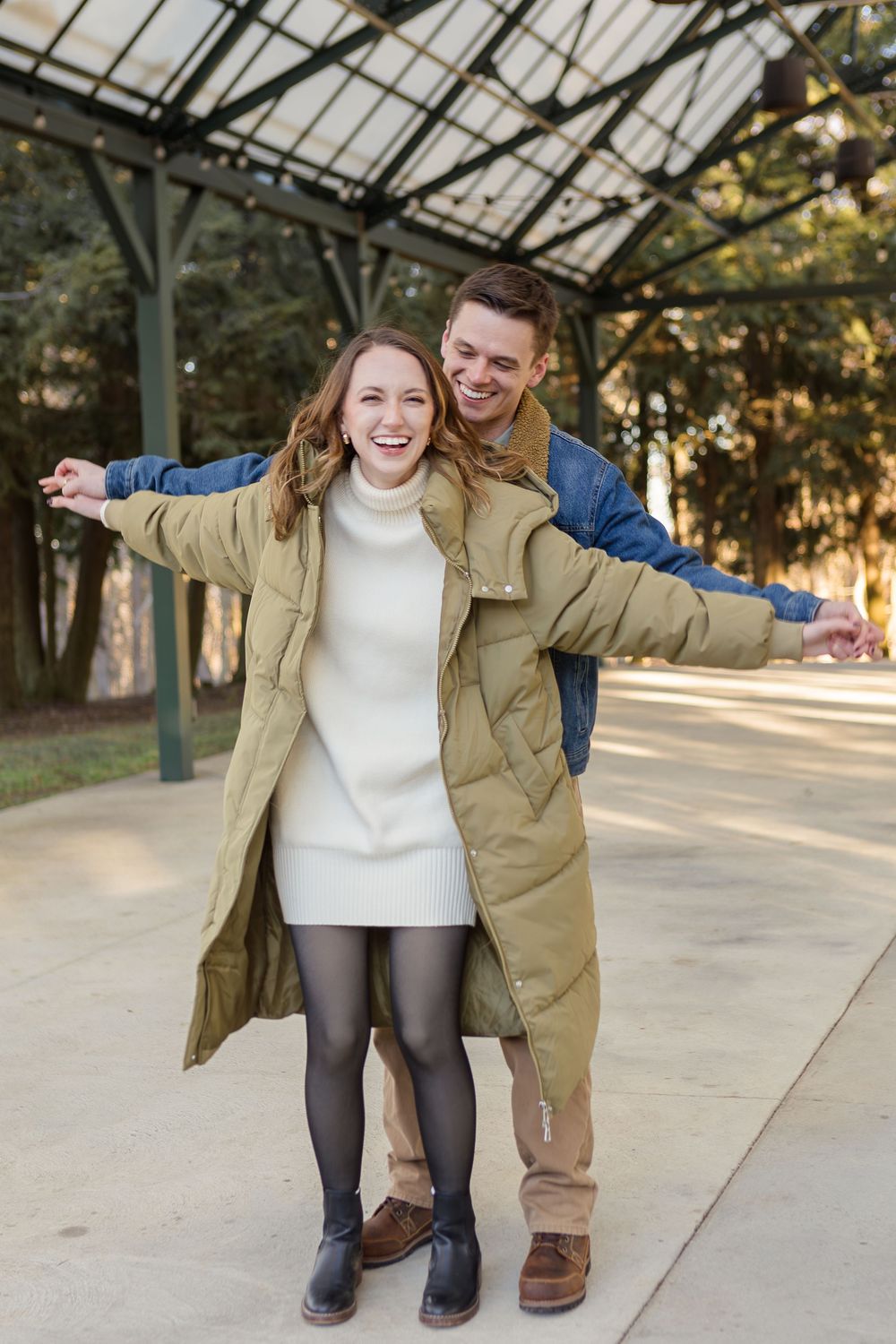 Smiling couple embracing outdoors under a covered pavilion, the woman in an olive puffer coat and white sweater dress with a man behind her.