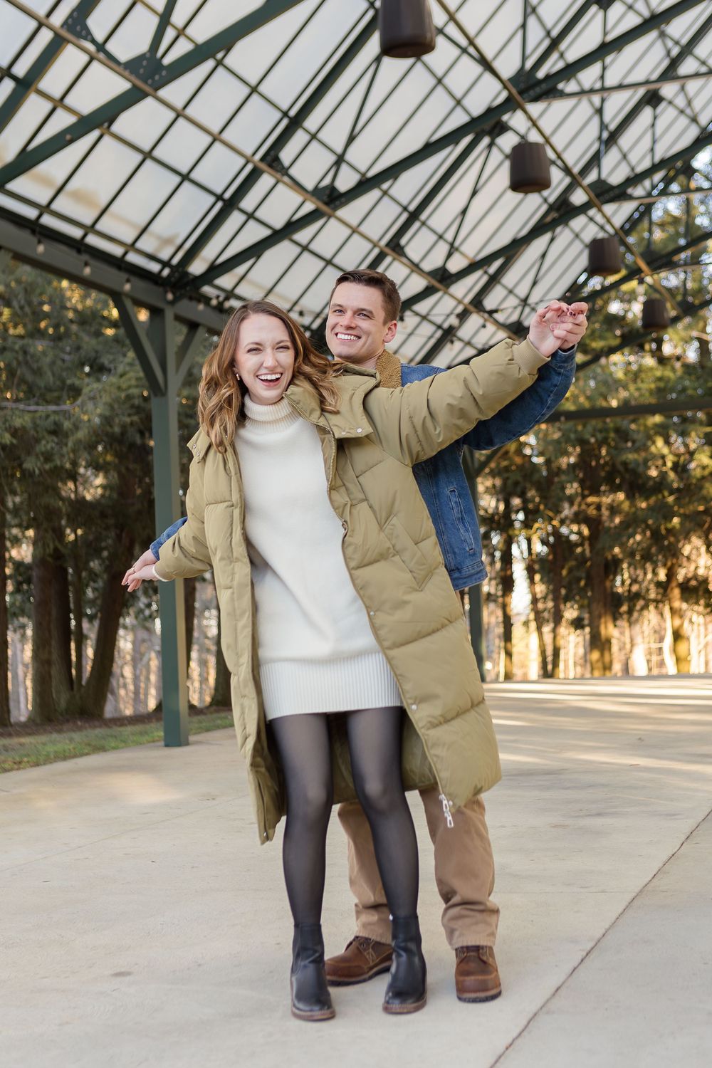 Smiling couple posing outdoors under a covered pavilion, the woman in an olive puffer coat with arms outstretched as the man embraces her fr