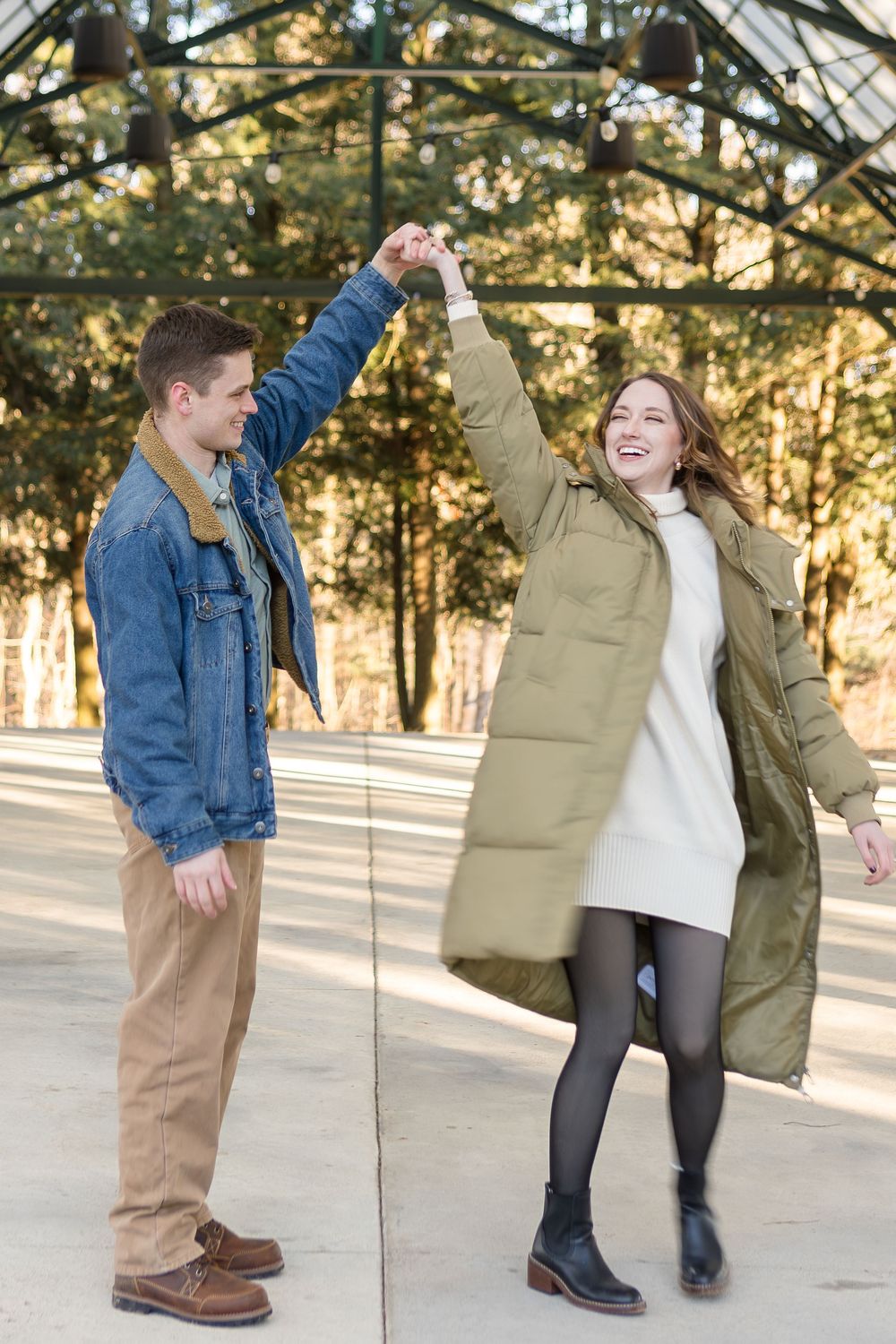 Smiling couple dancing outdoors as a man in a denim jacket twirls a woman in an olive puffer coat under a covered pavilion.