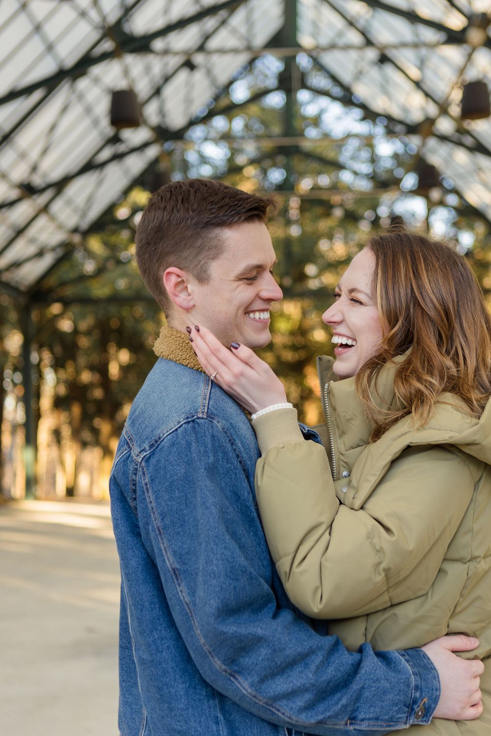 Smiling couple in denim jacket and olive puffer coat embrace and laugh together in a glass-roofed garden pavilion.
