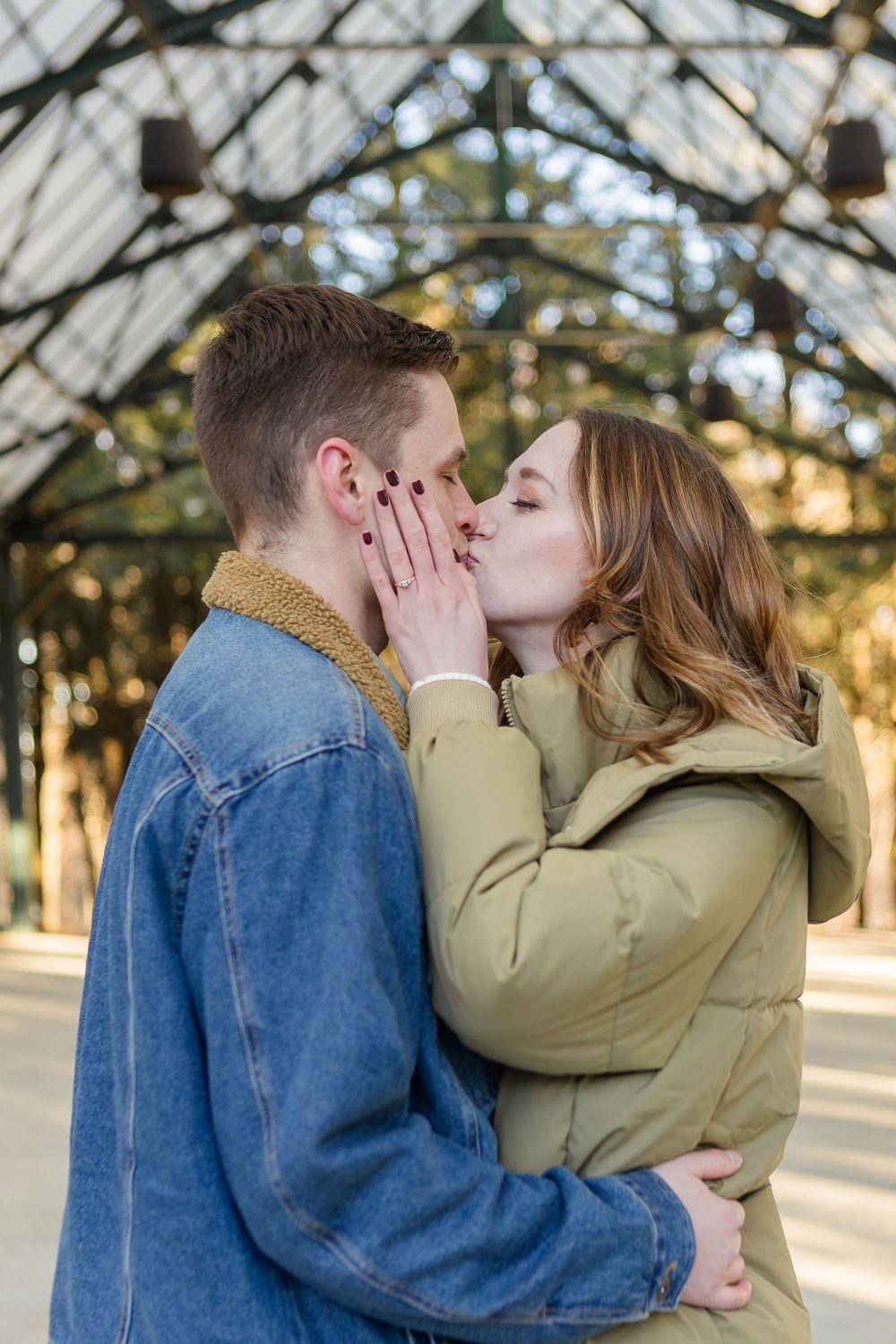 Couple in denim and olive puffer jackets share a kiss in a glass-roofed pavilion, her hand gently touching his lips.
