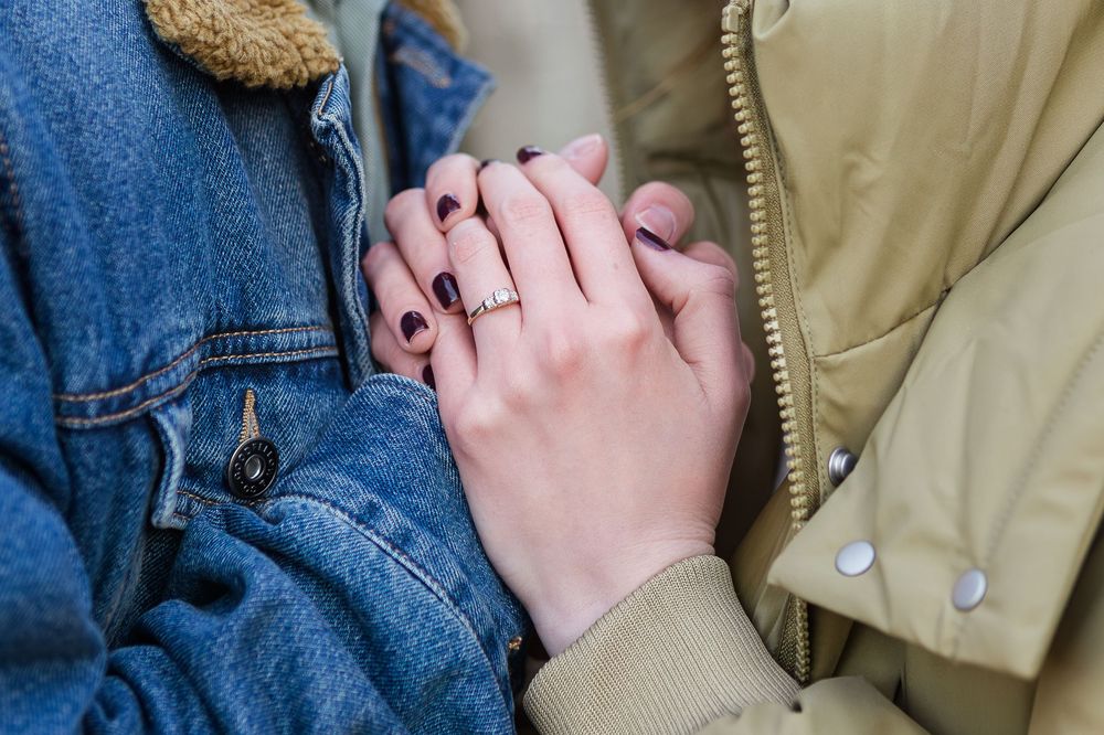 Close-up of two people holding hands, one wearing a denim jacket and the other a beige coat, with dark nail polish and a delicate ring.