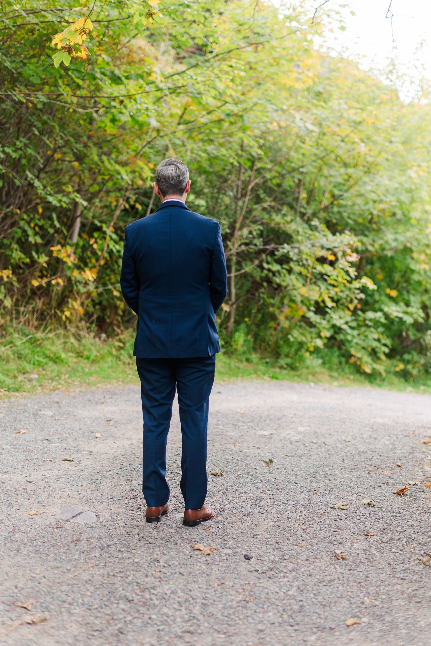 Man in a navy suit and brown leather shoes stands facing away on a gravel path surrounded by autumn trees.