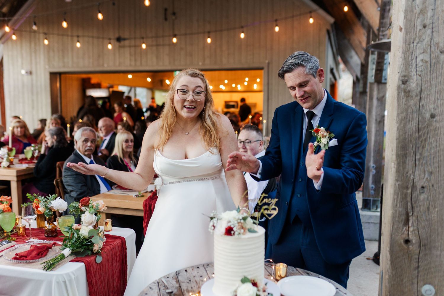 A bride in a white gown and a groom in a navy suit cut their wedding cake together as guests watch in a rustic barn venue.