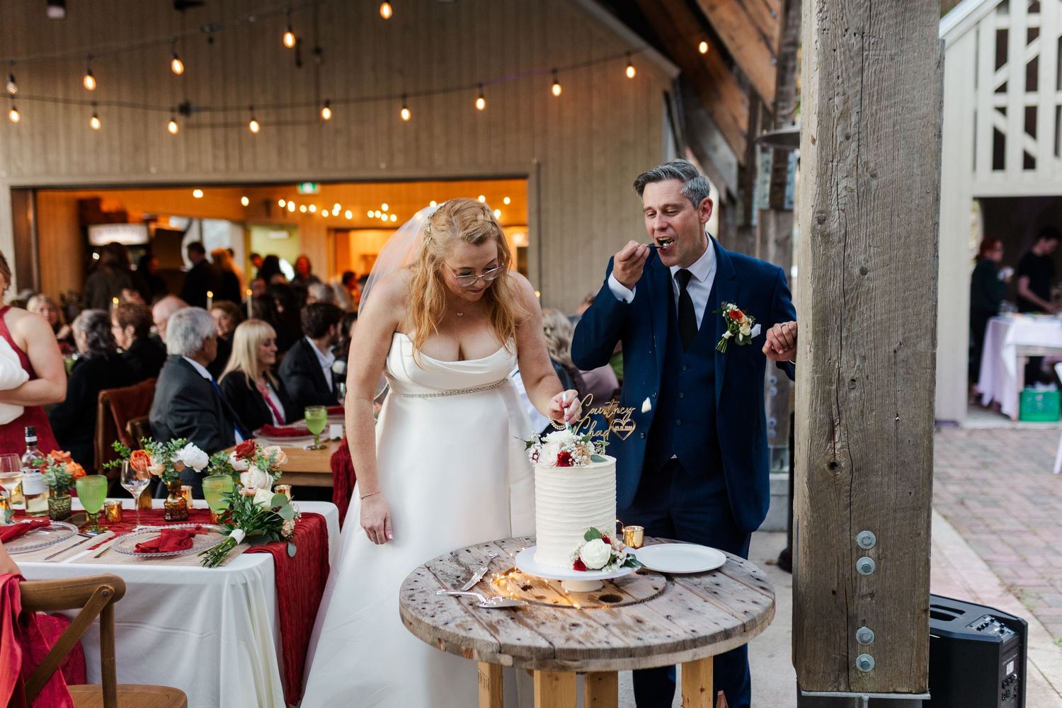 Bride in a white gown cuts a wedding cake on a rustic wooden table while the groom in a navy suit enjoys a bite at a barn reception.