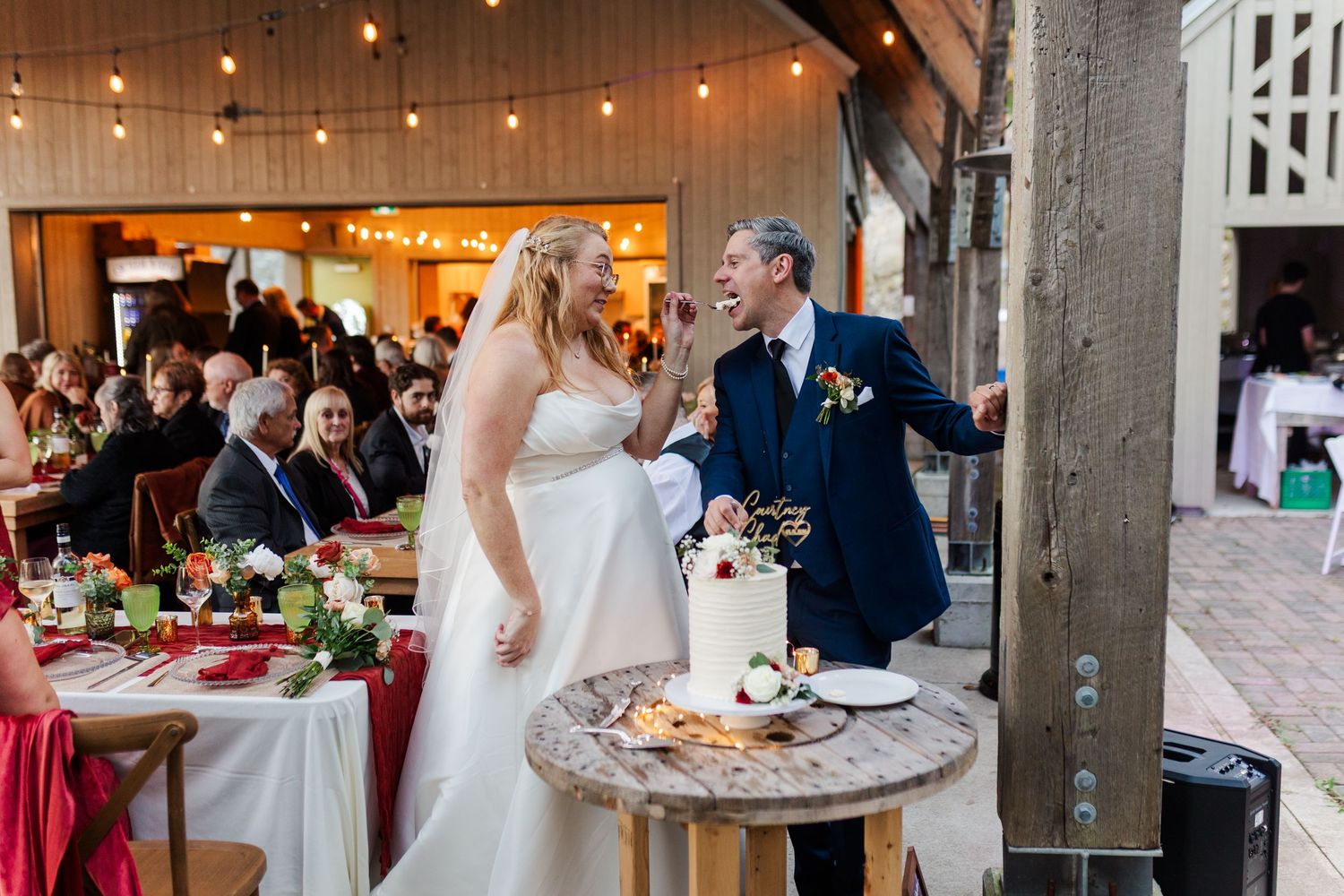 A bride laughs while feeding her groom a bite of wedding cake beside a rustic wooden table at a barn reception with string lights.