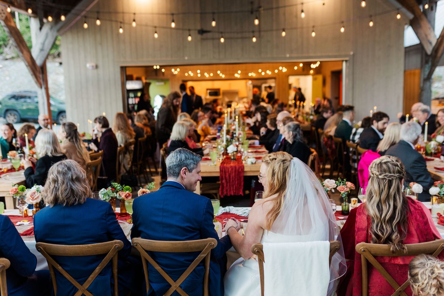 Bride in a white veil and groom seated at a long reception table with guests at a rustic outdoor wedding dinner under string lights.