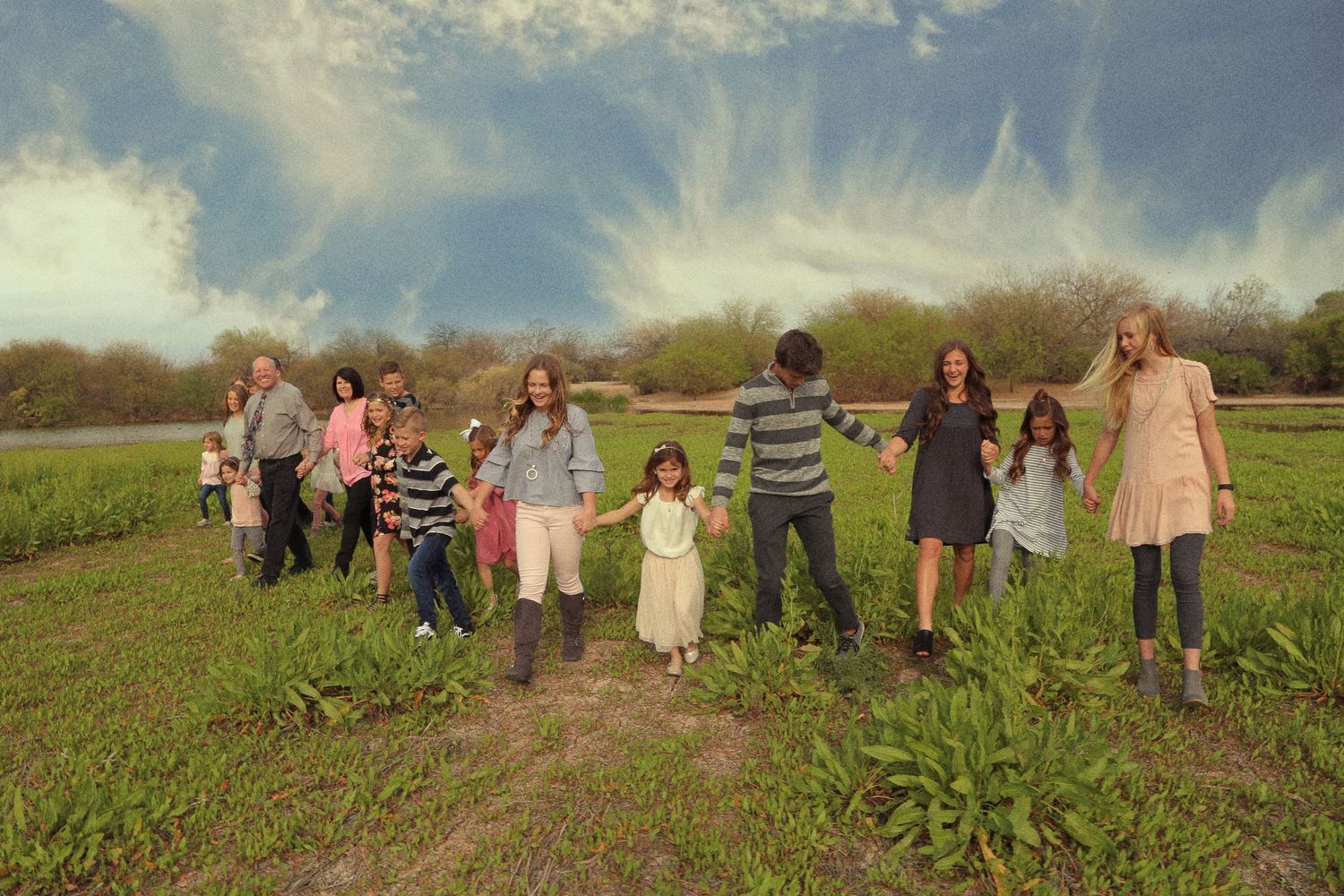 A large family holds hands and walks together through a green grassy field under a dramatic blue sky with wispy clouds.