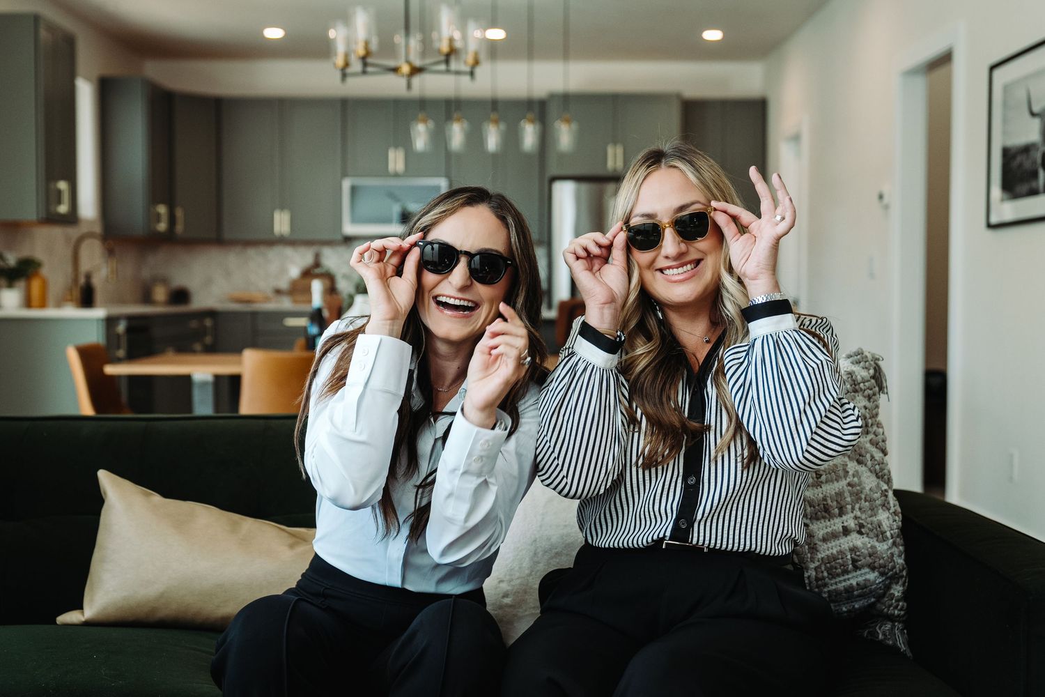 Two women sitting on a sofa wearing matching sunglasses and striped shirts, smiling in a modern living room.
