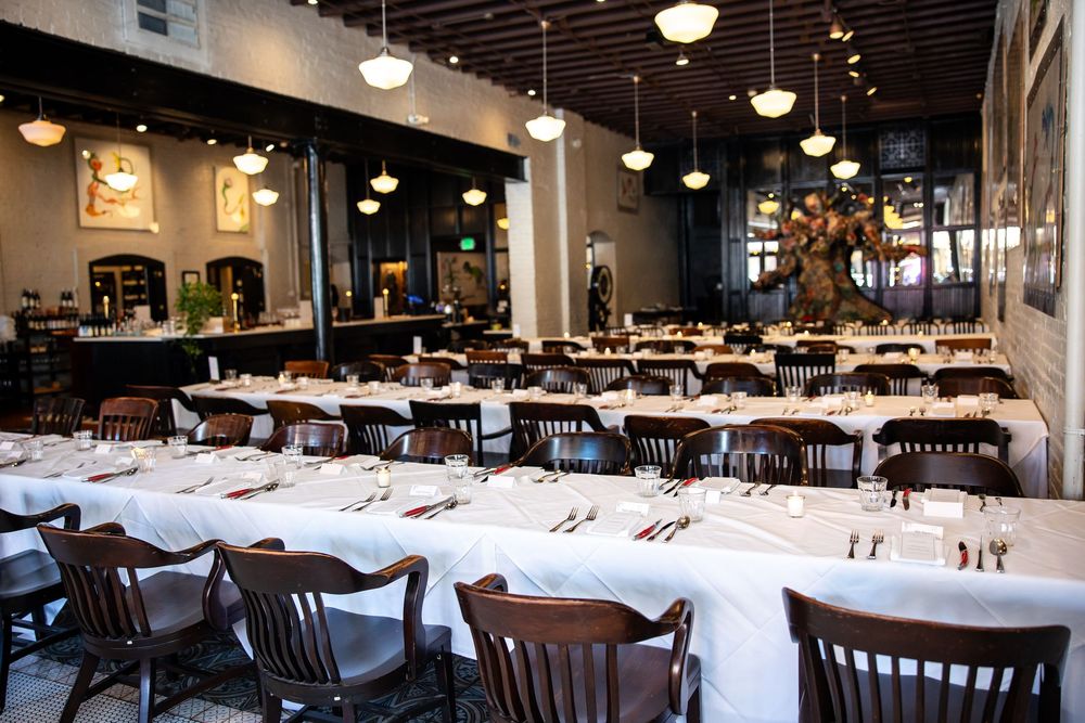 Elegant restaurant dining room with long white-clothed tables, dark wood chairs, and warm pendant lighting.