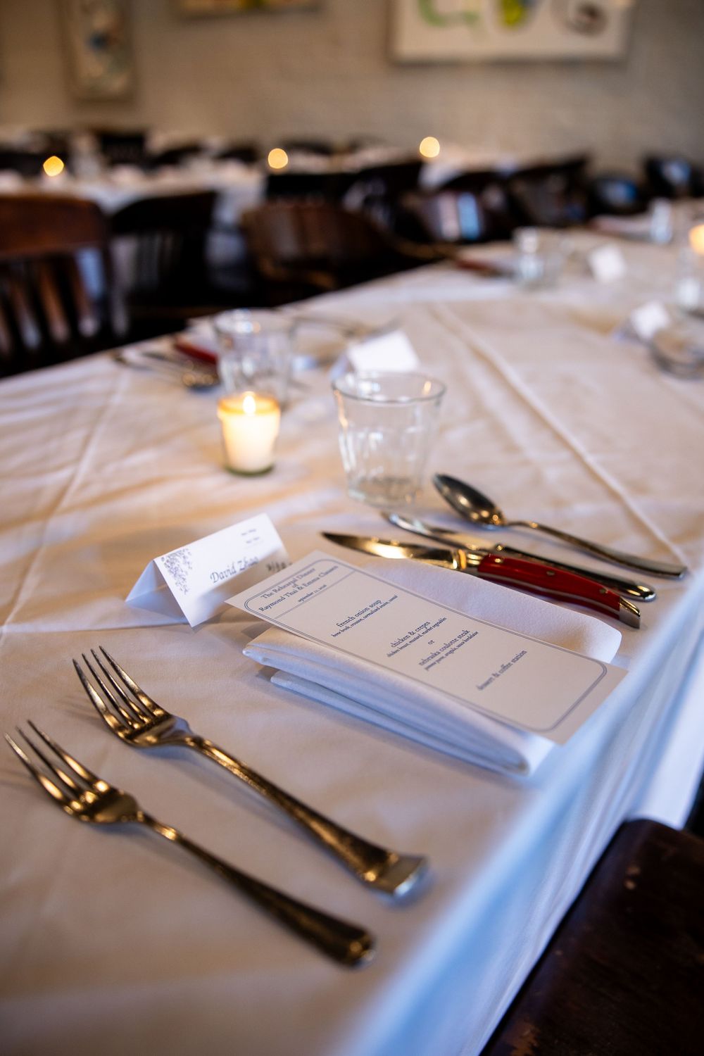 Elegant restaurant table setting with gold silverware, menu cards, name card, and lit candle on white linen tablecloth.