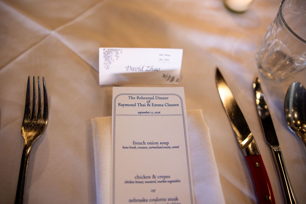 Elegant rehearsal dinner table setting with menu card, place card, fork, knife, and glassware on white linen tablecloth.