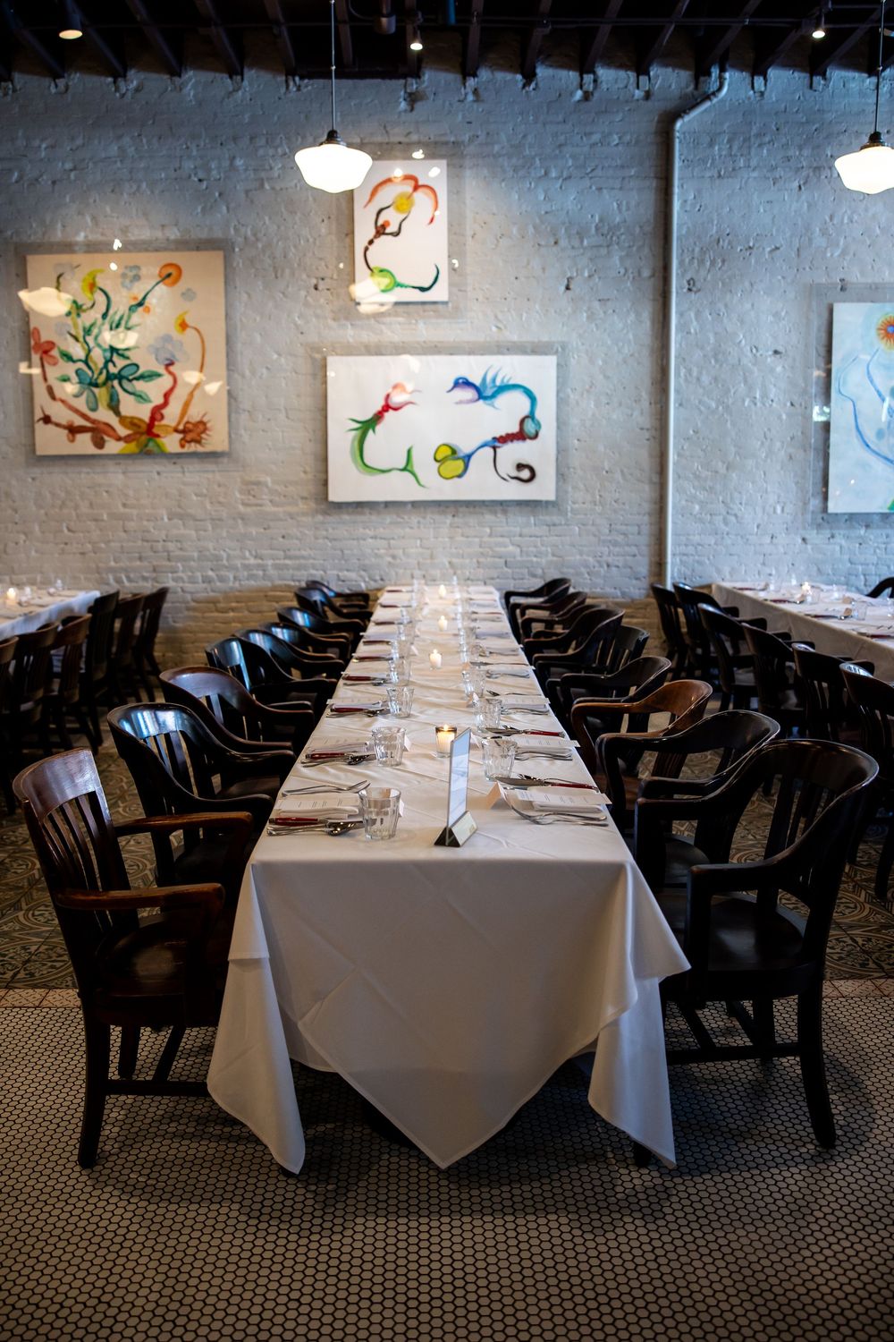 Elegant private dining room with long white-clothed table, dark wood chairs, white brick walls, and colorful abstract artwork.