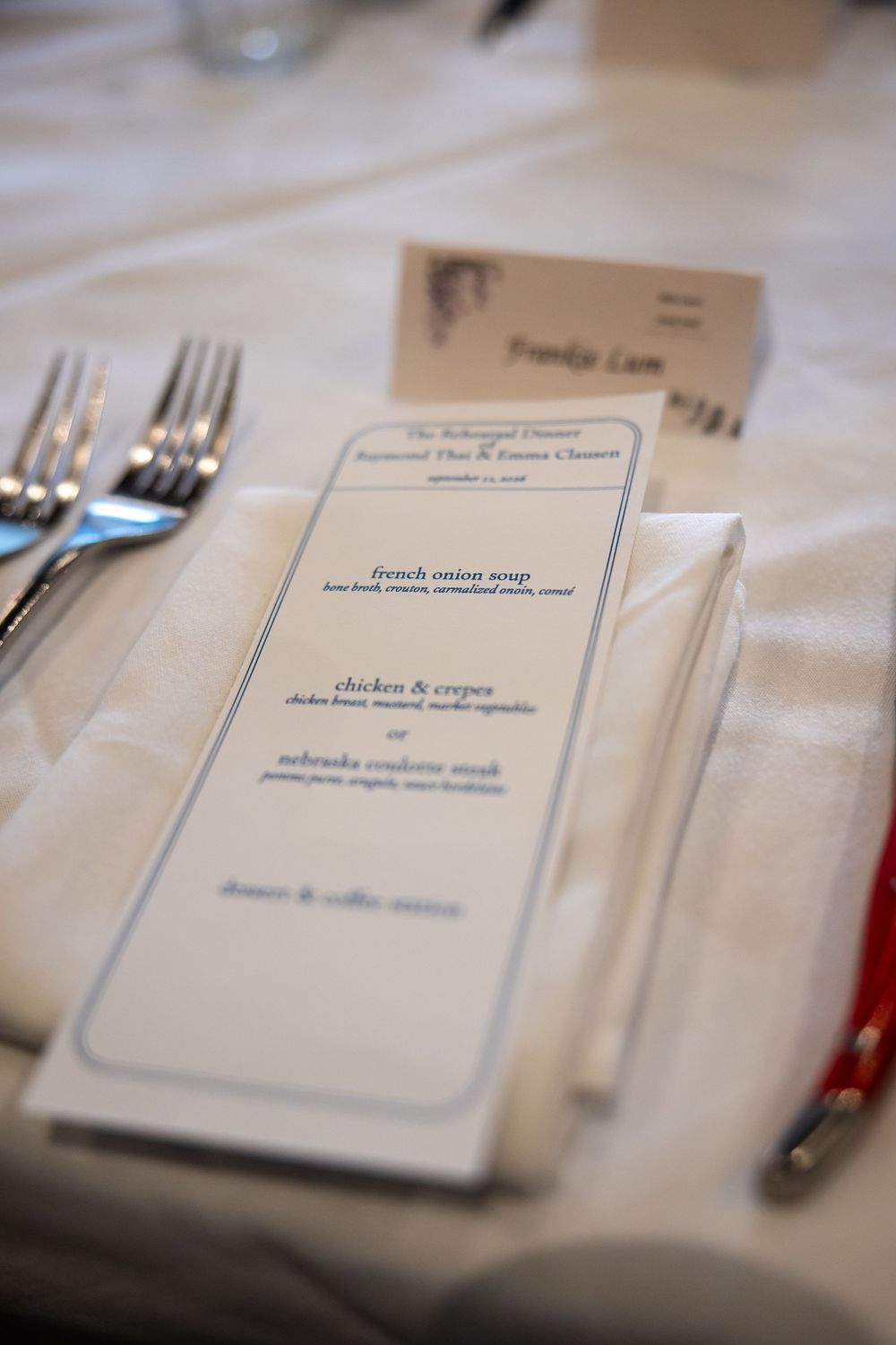 Elegant rehearsal dinner place setting with menu card, name card, white napkin, and silverware on a white tablecloth.