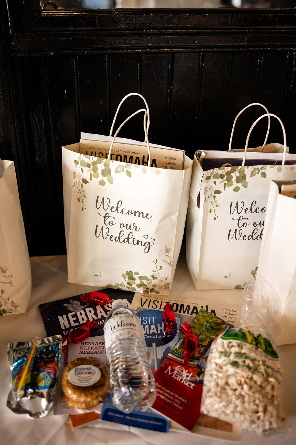 Wedding welcome bags with greenery design beside Nebraska souvenirs, snacks, and Visit Omaha brochures for guests.
