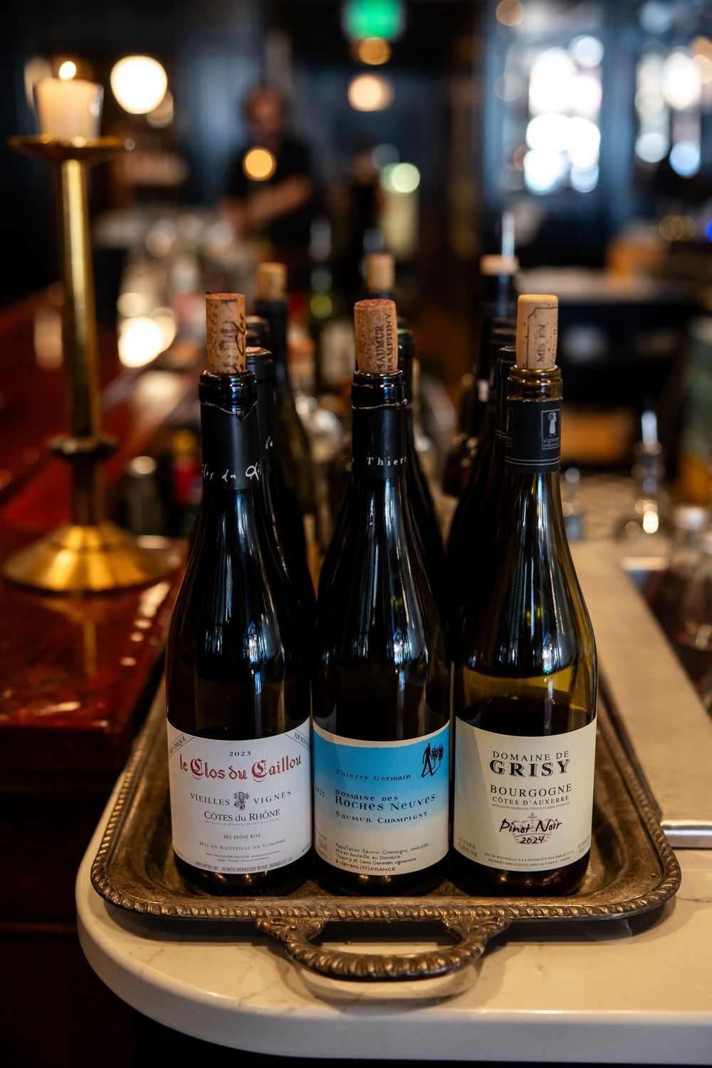 Three corked wine bottles on a silver tray at a bar, including Domaine de Grisy Bourgogne Pinot Noir.
